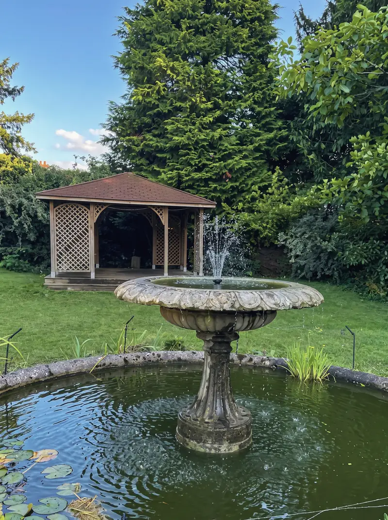 The timber arbour at the far end of the garden, behind the fountain and lily pond