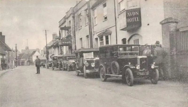 An old photograph of early motor cars parked in the street outside the hotel, under its hanging sign reading Avon Hotel