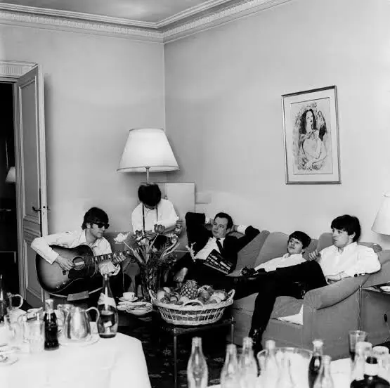 The Beatles in the Fountain Room in 1965: one plays an acoustic guitar while the others sit on a sofa beside a basket of fruit