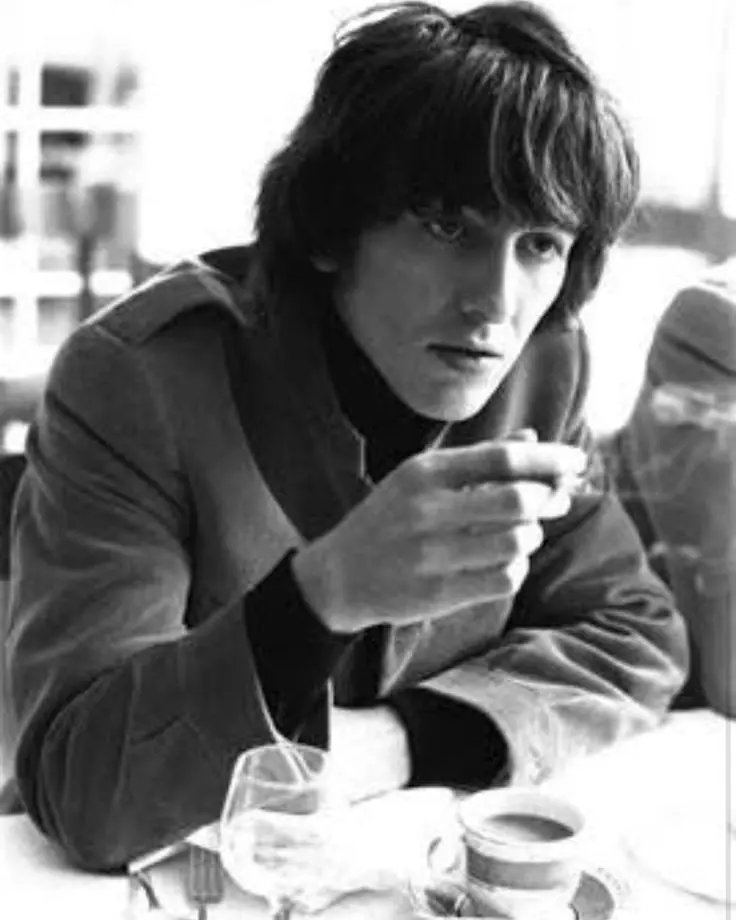 George Harrison at a table at the hotel in 1965, a cigarette in his hand beside a cup of coffee