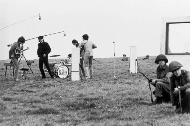 The Beatles filming Help! on Salisbury Plain in 1965: the band at their instruments in a grassy field under boom microphones, with soldiers crouching in the foreground