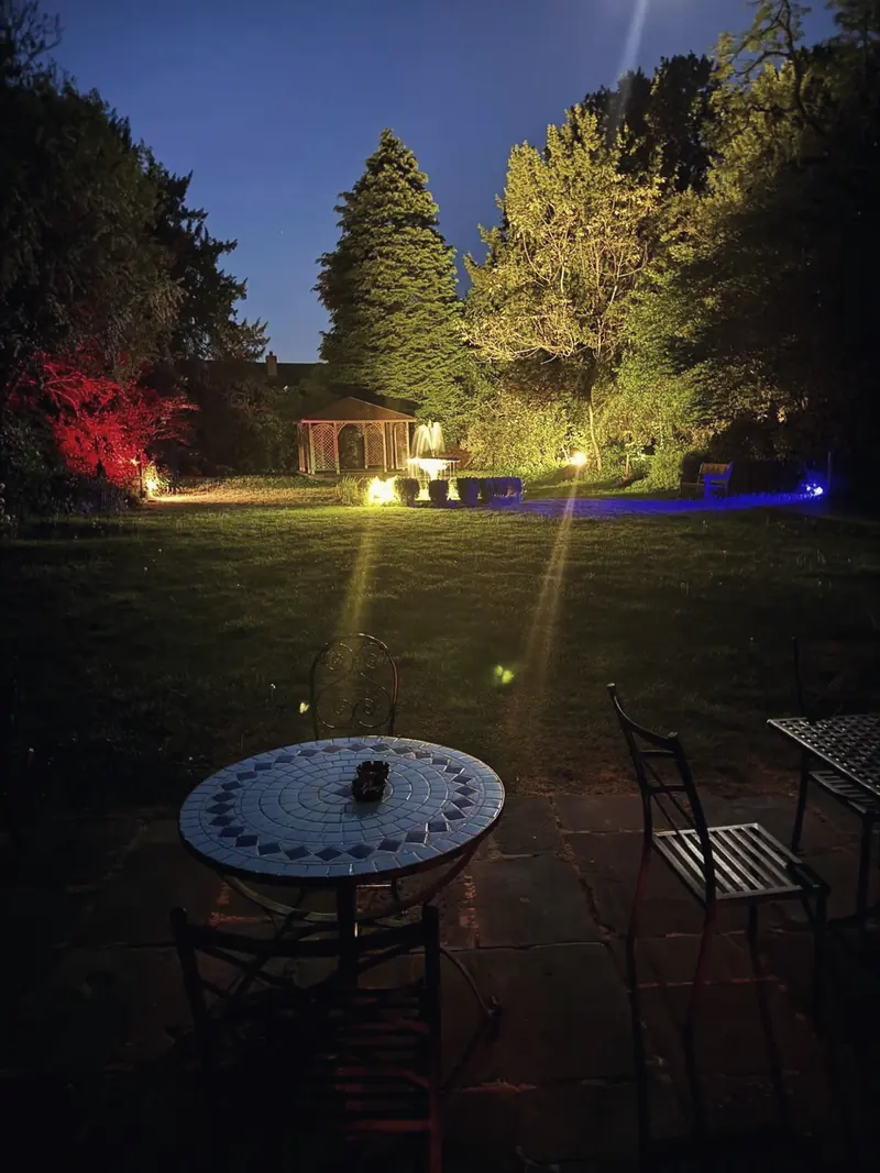The garden at dusk from the terrace, with a mosaic table in front and the arbour lit up beyond the lawn