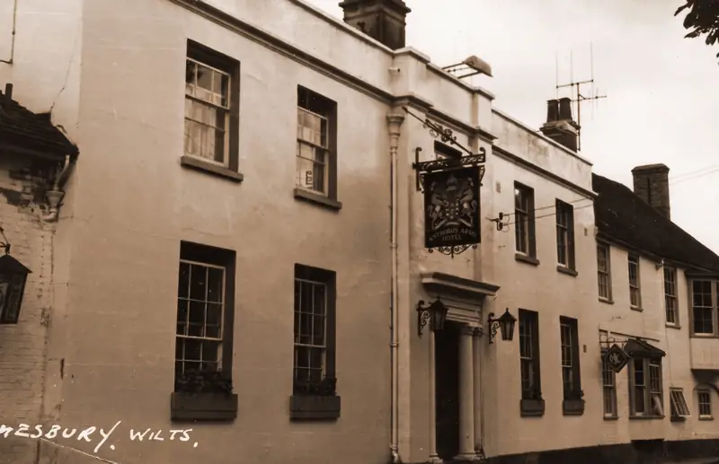 Old sepia postcard of the hotel's street front with a hanging inn sign, captioned Amesbury, Wilts