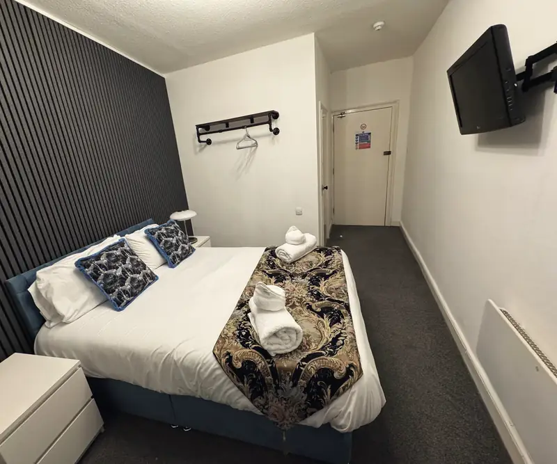 The Small Double Room: a double bed with a blue velvet base and a patterned throw against a dark slatted wall, with a wall-mounted TV and a hanging rail by the door